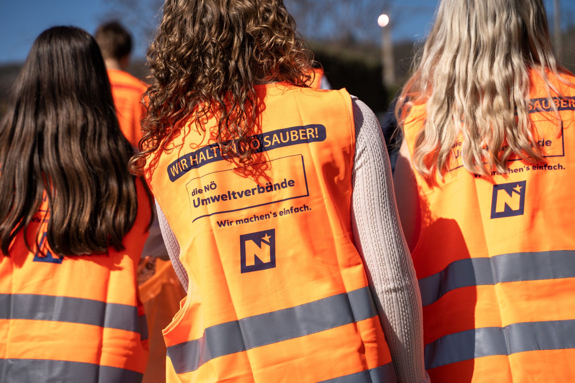 Auf dem Bild sind 3 Frauen von hinten zu sehen. Diese tragen eine orange Warnweste.