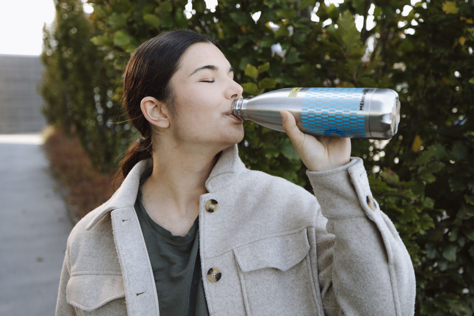 Ein jugendliches Mädchen mit brauen Haaren, einem grünen Oberteil sowie einer beigen Jacke, trinkt aus einer Edelstahlflasche. Auf der Edelstahlfalsche ist das Logo der NÖ Umweltverbände, unzählige kleine blaue Flaschen sowie die Aufschrift Mehrweg zu sehen.